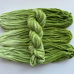 May include: Close-up of three skeins of green yarn, with a fourth skein of variegated green and white yarn in the center. The yarn is thick and chunky, ideal for knitting or crocheting projects.