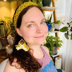 Pu&ograve; includere: Una donna con lunghi capelli castani che indossa una fascia per capelli in crochet gialla e orecchini a nappa gialli e bianchi. Indossa una canotta viola e blu.