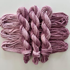 May include: Three twisted hanks of yarn arranged vertically over several horizontal loops of the same material. The yarn features a variegated color palette of soft mauve, dusty rose, and deep plum tones, creating a consistent gradient effect.