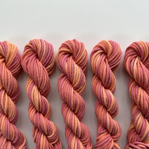 May include: Five skeins of yarn in a gradient of pink, peach, and orange hues. The yarn is tightly wound, showing a twisted rope-like texture. The background is a plain white surface, highlighting the vibrant colors of the yarn.