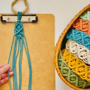 K&ouml;nnte beinhalten: Eine Sammlung von Makramee-Stirnb&auml;ndern mit komplizierten Knotenmustern in verschiedenen Farben wie Hellgr&uuml;n, Orange, Blaugr&uuml;n, Creme und Senfgelb, aufbewahrt in einem geflochtenen Korb neben einem blaugr&uuml;nen Stirnband an einer beigen Schlaufe.