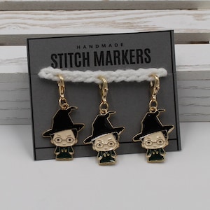 May include: Three gold stitch markers with black hats and green robes. The markers feature a cartoon character with glasses. The card reads "Handmade Stitch Markers".