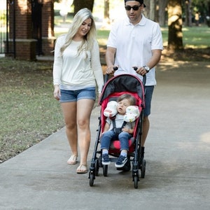 May include: A red stroller with a black frame and a child sitting inside. A woman and a man are walking behind the stroller on a paved path in a park.