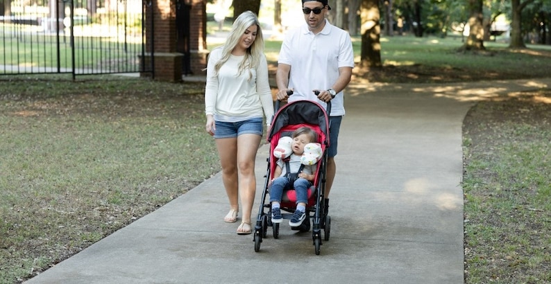 May include: A red stroller with a black frame and a child sitting inside. A woman and a man are walking behind the stroller on a paved path in a park.