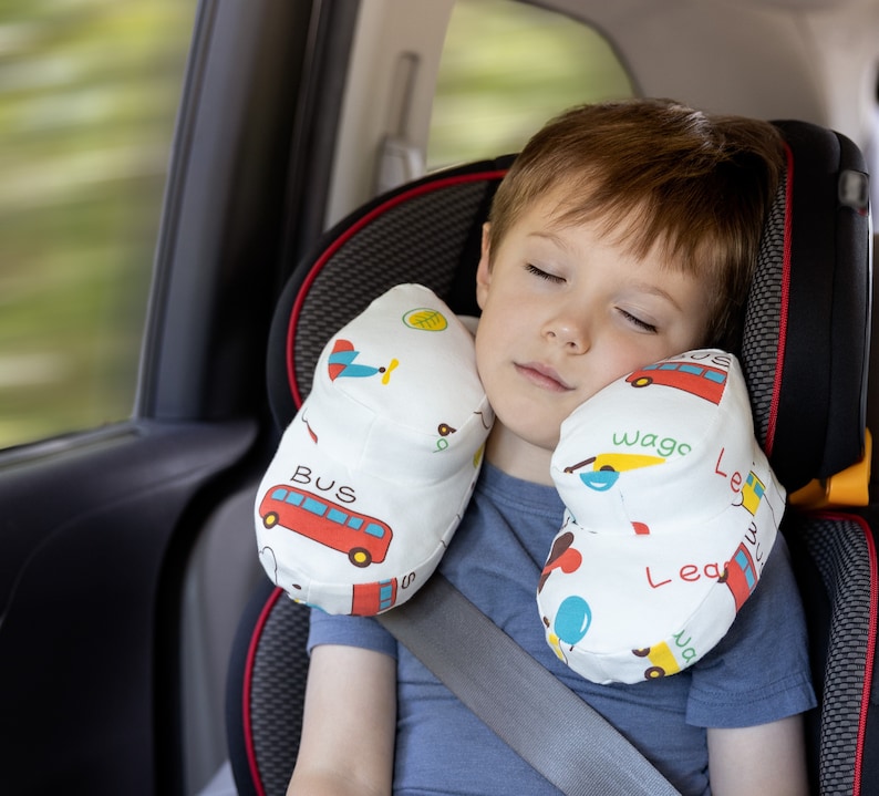 May include: A child sleeping in a car seat with a white and colorful travel pillow with a bus, airplane, and other designs. The pillow has the words "Bus", "Wago", "Lea", and "Lea" printed on it.