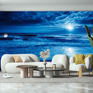 May include: A wall mural featuring a dark blue night ocean scene with a bright moon and rolling waves. The room includes a curved white sofa, a round coffee table with a speckled base, a white armchair with a yellow pillow, and a tall green plant.