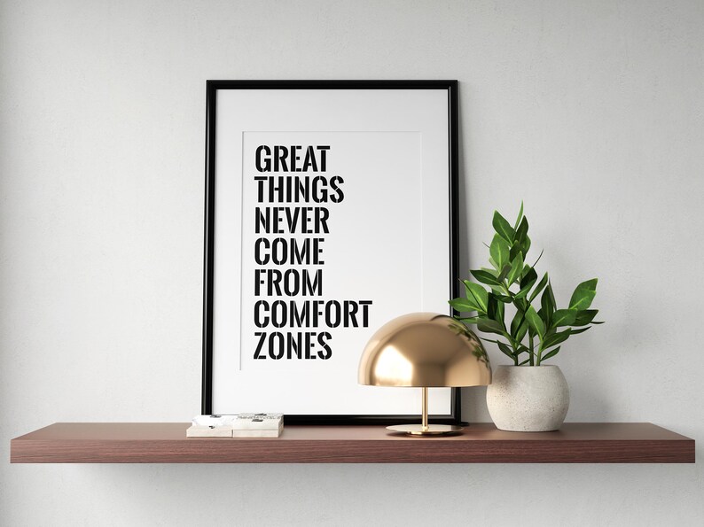 May include: A black framed print with the text "GREAT THINGS NEVER COME FROM COMFORT ZONES" on a white background. A gold lamp and a potted green plant are on a brown shelf.