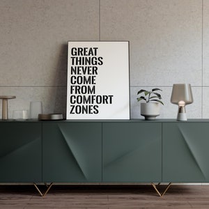 May include: A green cabinet with gold legs and three doors. The cabinet is decorated with a geometric pattern. A black and white poster with the text "Great things never come from comfort zones" is displayed above the cabinet. A small plant in a white pot and a gray table lamp are on the cabinet.