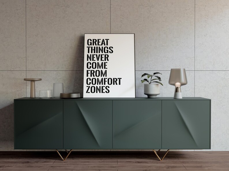 May include: A green cabinet with gold legs and three doors. The cabinet is decorated with a geometric pattern. A black and white poster with the text "Great things never come from comfort zones" is displayed above the cabinet. A small plant in a white pot and a gray table lamp are on the cabinet.