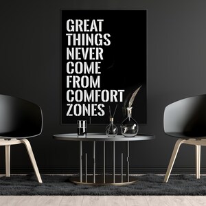 May include: Black and white motivational quote poster with the text "GREAT THINGS NEVER COME FROM COMFORT ZONES" hanging on a black wall with two black chairs and a black coffee table with decorative items.