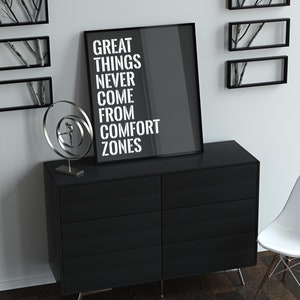 May include: A black and white motivational poster with the text "Great things never come from comfort zones". The poster is framed and hanging on a white wall above a black dresser with a white chair to the right.