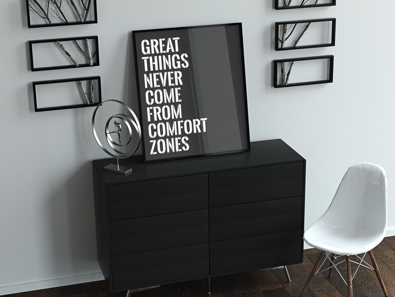 May include: A black and white motivational poster with the text "Great things never come from comfort zones". The poster is framed and hanging on a white wall above a black dresser with a white chair to the right.