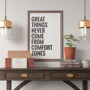 May include: A framed print with the text "Great things never come from comfort zones" hanging on a white wall above a wooden console table. The table has two drawers with gold-toned pulls. There are two gold pendant lights hanging above the table, a potted plant, a stack of books, and a small wooden box with a label on top.