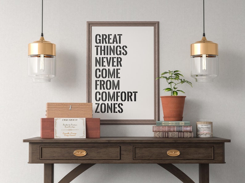 May include: A framed print with the text "Great things never come from comfort zones" hanging on a white wall above a wooden console table. The table has two drawers with gold-toned pulls. There are two gold pendant lights hanging above the table, a potted plant, a stack of books, and a small wooden box with a label on top.