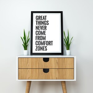 May include: A black framed print with the text "GREAT THINGS NEVER COME FROM COMFORT ZONES" on a white background. The print is sitting on a wooden nightstand with two drawers and black handles. There are two small green plants in white pots on either side of the print.