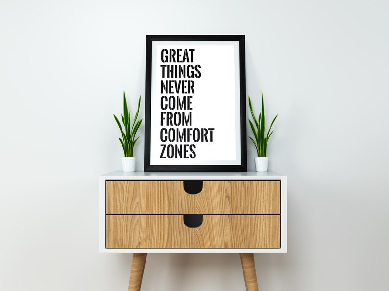 May include: A black framed print with the text "GREAT THINGS NEVER COME FROM COMFORT ZONES" on a white background. The print is sitting on a wooden nightstand with two drawers and black handles. There are two small green plants in white pots on either side of the print.