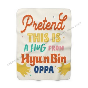 Pu&ograve; includere: Una coperta color crema con il testo "Pretend this is a hug from Hyun Bin Oppa" in lettere colorate e stilizzate. Il testo &egrave; circondato da piccole stelle e due mani gialle che si tendono per un abbraccio.