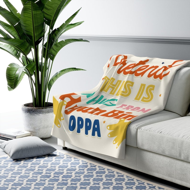 Pu&ograve; includere: Una coperta bianca con il testo "Pretend this is from your bday Oppa" in lettere colorate e giocose.
