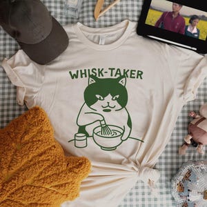 May include: Cream-colored t-shirt with a green graphic of a cat whisking in a bowl, with the text "WHISK-TAKER" above. The shirt is tied in a knot at the bottom. A brown baseball cap and a yellow pillow are in the background.
