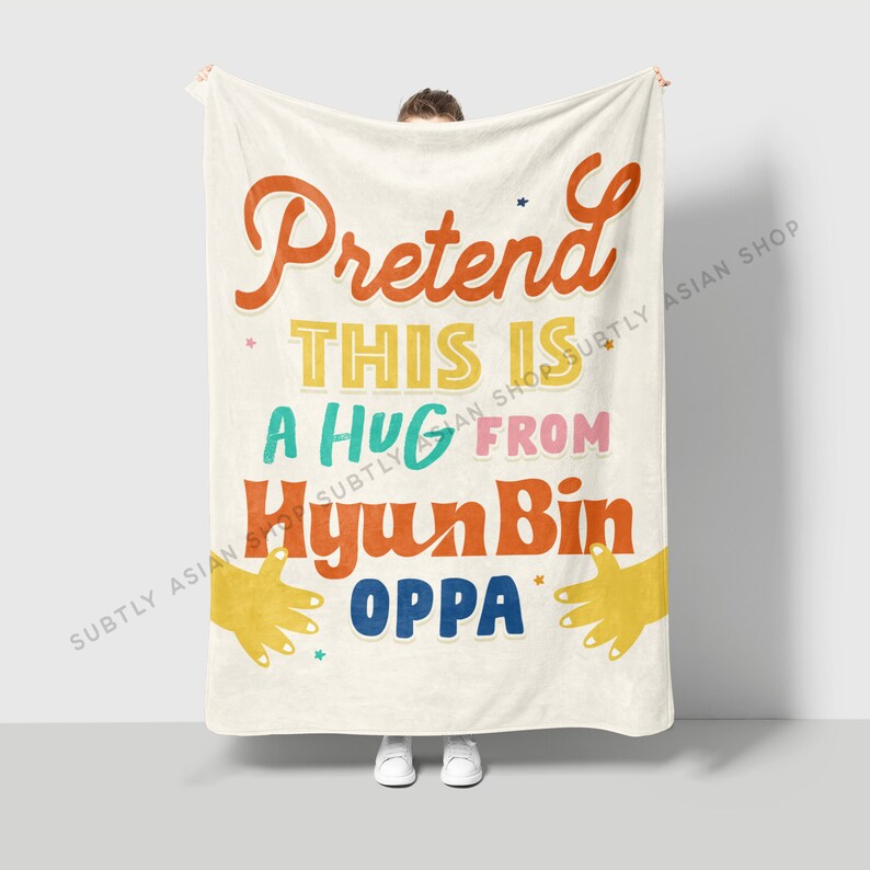 Pu&ograve; includere: Una coperta in pile bianca con il testo "Pretend This Is A Hug From Hyun Bin Oppa" in lettere colorate. Il testo &egrave; circondato da contorni di mani gialle.