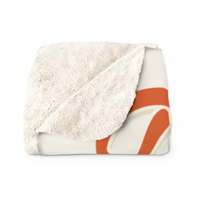 Pu&ograve; includere: Una morbida coperta in pile, di colore bianco e crema, con un design arancione e crema. La coperta &egrave; piegata a met&agrave; e il design &egrave; visibile sul lato anteriore.