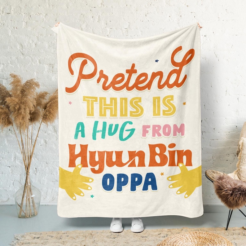 Pu&ograve; includere: Una coperta in pile bianco con il testo "Pretend this is a hug from Hyun Bin Oppa" in lettere colorate in stile retr&ograve;. Due mani gialle sono su entrambi i lati del testo, come se stessero dando un abbraccio.