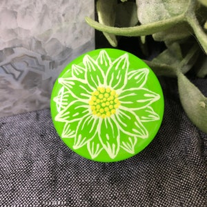 May include: A green painted rock with a white daisy flower design. The daisy has a yellow center with white petals.