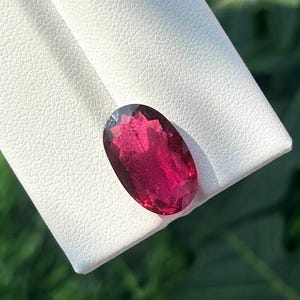 May include: An oval-cut, deep red gemstone with facets, resting on a white textured surface. The gemstone's color is a rich, saturated hue, and it appears to be polished. The background is blurred, suggesting a natural setting.