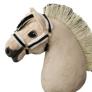 May include: A plush horse head toy in a light cream color with a long, thick mane made of cream and black yarn. The horse features a black and white halter with soft white faux fur padding, dark eyes, and a dark gray muzzle and nostril area.
