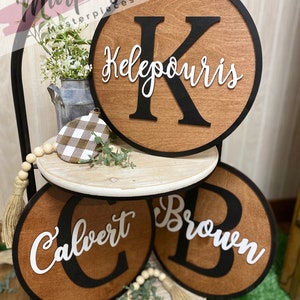 May include: Three wooden round signs with black borders and white lettering. The top sign says "K  Kelepouris", the middle sign says "C Calvert", and the bottom sign says "Brown". The signs are on a tiered stand with a white background.