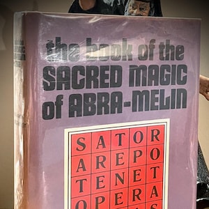 May include: A hardcover book titled "The Book of the Sacred Magic of Abra-Melin" with a purple cover. The cover features the title in large black letters and a square with the words "SATOR AREPO TENET OPERA ROTAS".