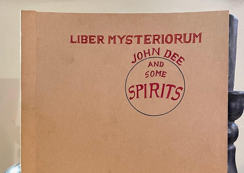 May include: A book cover with the title "Liber Mysteriorum" in red letters. Below, a circular design reads "John Dee and Some Spirits" in red. The cover is a light brown color.