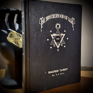 Op de afbeelding: Een vintage zwart boek getiteld "The Brotherhood of Light: Sacred Tarot" door C.C. Zain. De omslag heeft sierlijke witte letters en een stersymbool. Het boek wordt vastgehouden door een zwart handbeeld.