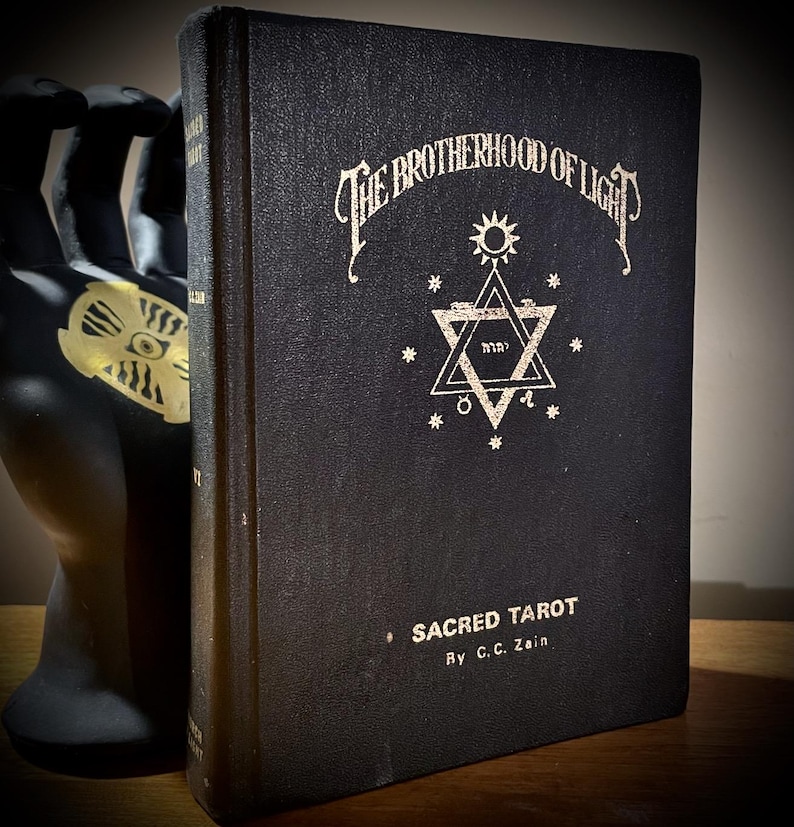 Op de afbeelding: Een vintage zwart boek getiteld "The Brotherhood of Light: Sacred Tarot" door C.C. Zain. De omslag heeft sierlijke witte letters en een stersymbool. Het boek wordt vastgehouden door een zwart handbeeld.