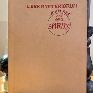 May include: A vintage-style book with a tan cover. The title "Liber Mysteriorum" is printed in red, along with "John Dee and Some Spirits" in a circular design. The book is displayed upright.