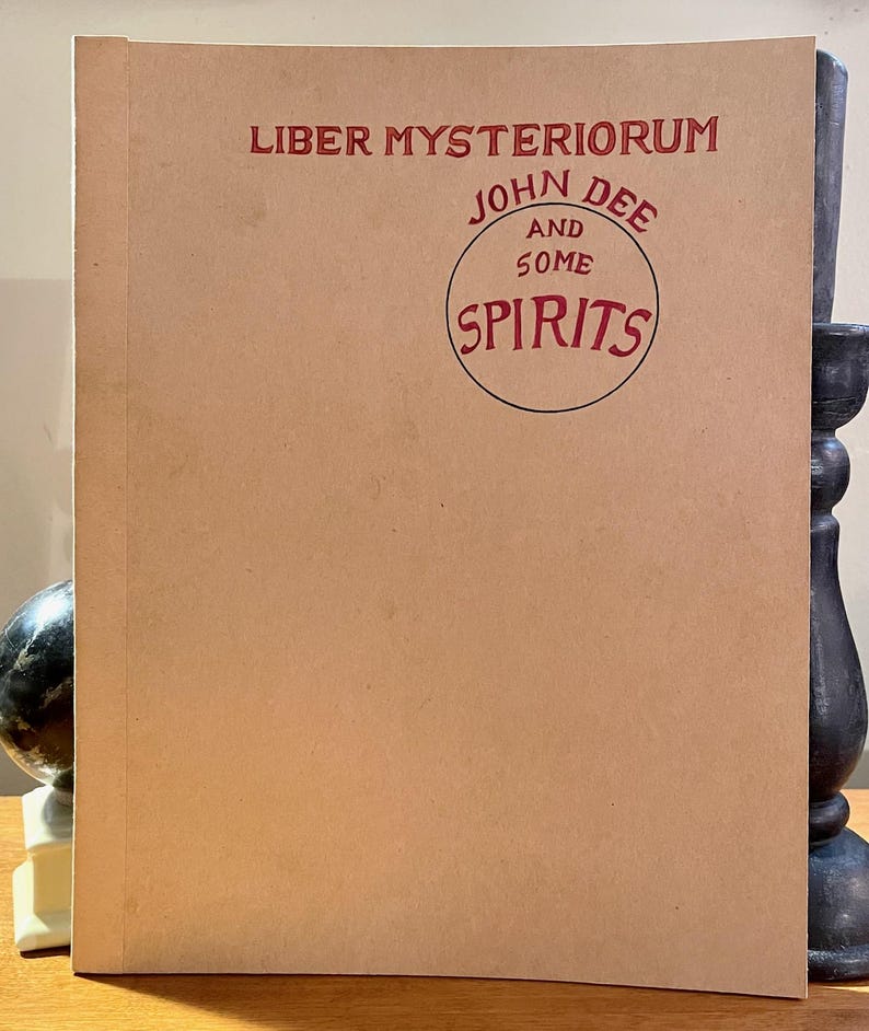 May include: A vintage-style book with a tan cover. The title "Liber Mysteriorum" is printed in red, along with "John Dee and Some Spirits" in a circular design. The book is displayed upright.