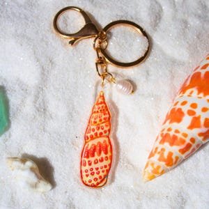 May include: A gold-colored keychain with a seashell charm. The charm is a vibrant orange and red seashell design with a pearl accent. The keychain is set against a white, sandy background with other seashells.