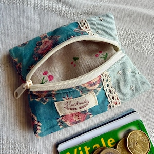 May include: A small zippered pouch made of teal floral fabric and light blue denim, featuring a cream lace trim, a white zipper, and a beige label with the word handmade. The interior lining is beige with a small red cherry print pattern.