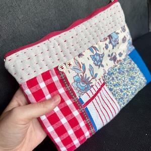 Peut inclure: Une pochette rectangulaire à fermeture éclair en tissu patchwork, composée d'une section à carreaux vichy rouges et blancs, d'un imprimé floral bleu sur fond crème et d'un panneau crème avec des surpiqûres rouges. Deux petits boutons blancs.