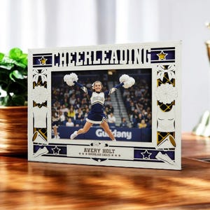 May include: A white picture frame with the word "CHEERLEADING" cut out at the top. The frame surrounds a photo of a cheerleader jumping in the air. The frame has blue and gold accents and the name "AVERY HOLT" is at the bottom.
