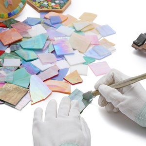 Pu&ograve; includere: Una persona con guanti bianchi usa un tagliavetro per tagliare un pezzo di vetro iridescente. Il vetro &egrave; circondato da altri pezzi di vetro di vari colori e forme. &Egrave; anche visibile un paio di tagliavetro sullo sfondo.