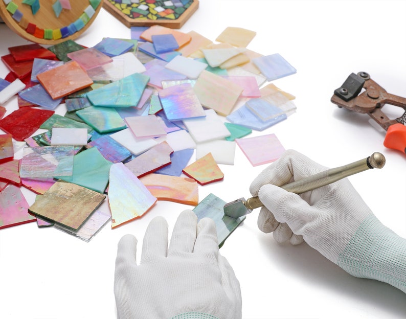 Pu&ograve; includere: Una persona con guanti bianchi usa un tagliavetro per tagliare un pezzo di vetro iridescente. Il vetro &egrave; circondato da altri pezzi di vetro di vari colori e forme. &Egrave; anche visibile un paio di tagliavetro sullo sfondo.