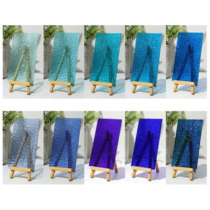 May include: A collection of ten rectangular glass panels in various shades of blue, green, and purple. Each panel has a textured surface and is displayed on a small wooden easel. The panels are arranged in two rows of five, showcasing a range of colors.