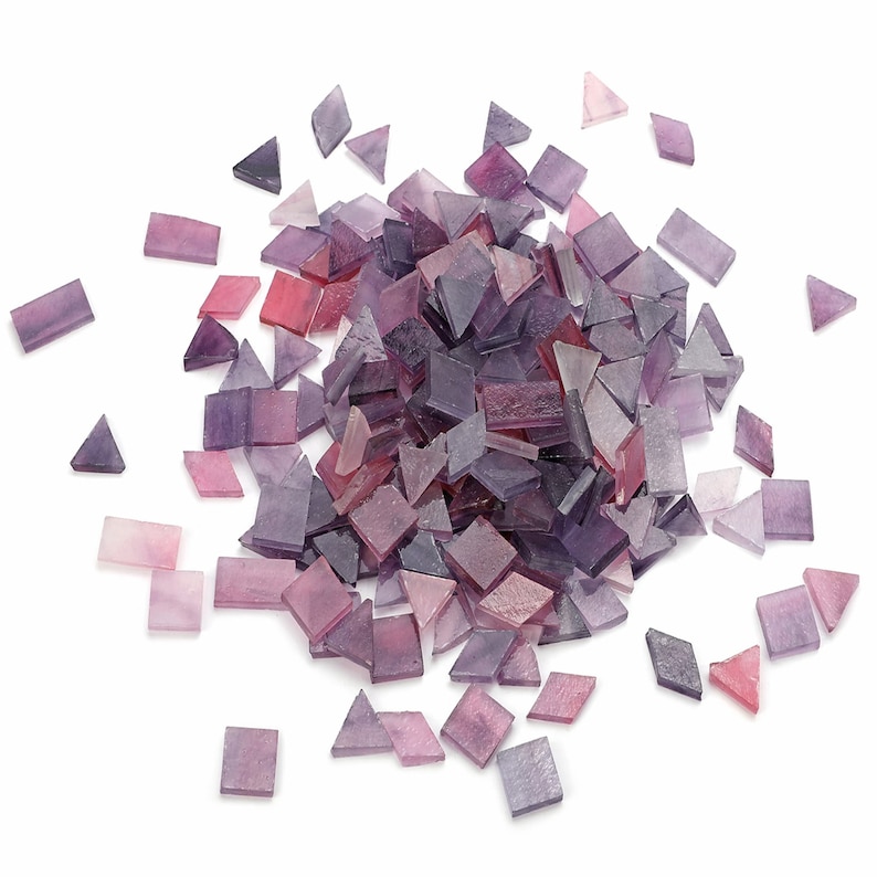 May include: A large pile of purple and pink glass mosaic tiles, scattered on a white background. The tiles are various shapes and sizes, including squares and triangles.