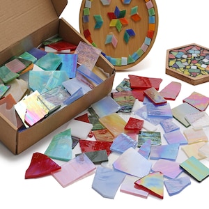 May include: A box filled with colorful glass mosaic tiles, some iridescent, with a few tiles scattered on a white surface. The tiles are various shapes and sizes, perfect for crafting projects.