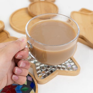 Op de afbeelding: Een glazen mok gevuld met een lichtbruine drank, rustend op een houten onderzetter met een stervormig moza&iuml;ekontwerp. De onderzetter is omringd door andere houten onderzetters in verschillende vormen.