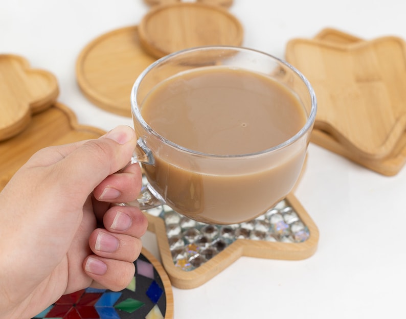 Op de afbeelding: Een glazen mok gevuld met een lichtbruine drank, rustend op een houten onderzetter met een stervormig moza&iuml;ekontwerp. De onderzetter is omringd door andere houten onderzetters in verschillende vormen.