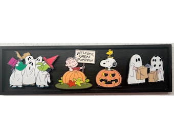 Op de afbeelding: Een zwart omkaderde Halloween-decoratie met Peanuts-personages. De scène bevat geesten, heksen, Linus met een bord met de tekst "Welcome Great Pumpkin", Snoopy op een pompoen en meer geesten. De personages zijn in een kleurrijke cartoonstijl.