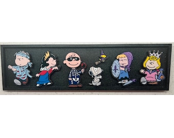 Op de afbeelding: Een rechthoekig zwart displaybord met zes Peanuts-emaille pins in Halloween-kostuums: een mummie, een koningin, een skelet, een mummiehond, een ridder met een schild en een prinses met een pompoentas, plus een kleine heksenvogel.