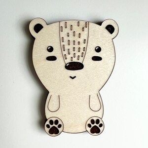 May include: A wooden cutout of a bear with a brown nose and black eyes. The bear has a pattern of small circles on its back and paw prints on its feet.